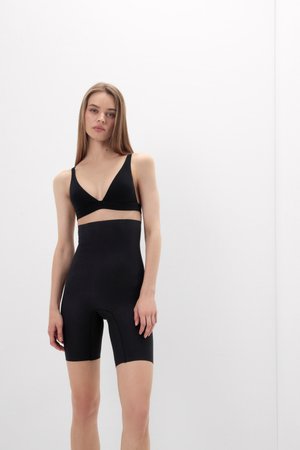 Femme portant un soutien-gorge noir et un short gainant noir taille haute, debout devant un fond blanc uni.
