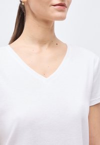 Γυναίκα που φοράει λευκό T-shirt με V λαιμόκοψη και χρυσό κρίκο στο αυτί, δείχνει τον λαιμό και το κάτω μέρος του προσώπου με ουδέτερη έκφραση σε φωτεινό φόντο.