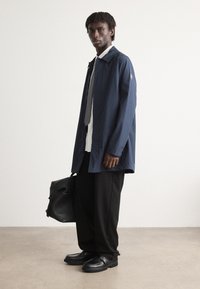 Mornarski vodootporni kaput, dugi rukavi, rašireni ovratnik. Bijela košulja, siva kravata, crne široke hlače, crne cipele. Crna kožna torba u ruci.