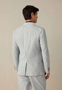 Helehall tekstuuriga ülikonnajakk, millel on üks tagaõmblus, kaks nuppu ja pikad varrukad, vaadatuna tagant neutraalse tausta ees.