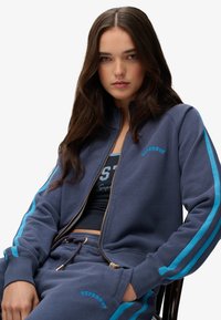 Sudadera con cremallera azul marino con rayas turquesas, tela suave y cremallera dorada, combinada con pantalones deportivos a juego que cuentan con el logo de la marca y cordón ajustable.