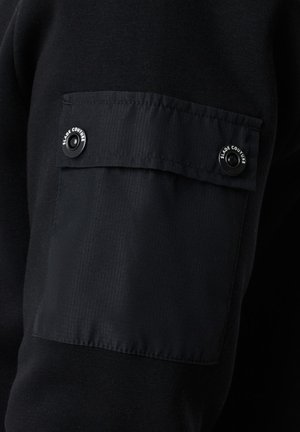 Musta dressipluusi kangas, millel on tekstuuriga overlay-patch varrukal, millel on kaks nööpi, millele on embossitud 'SLADE COUTURE'.