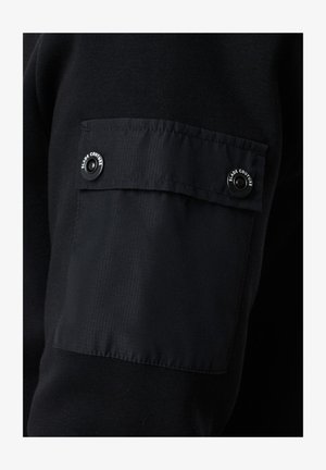 Musta dressipluusi kangas, millel on tekstuuriga overlay-patch varrukal, millel on kaks nööpi, millele on embossitud 'SLADE COUTURE'.