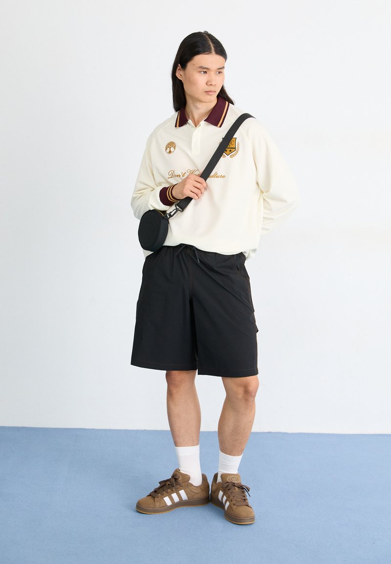 Polo de manga larga color blanco roto con un cuello burdeos, pantalones cortos negros y zapatillas marrones con rayas blancas. Bandolera negra llevada en el hombro.