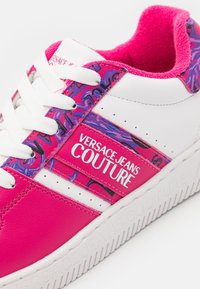 Baskets blanches et roses Versace Jeans Couture avec des accents à motifs violets et des lacets blancs, affichant le logo de la marque sur le panneau latéral.