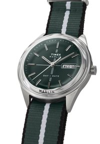Het Timex Marlin horloge heeft een groene plaat, zilveren uurmarkers en een dag/datumaanduiding; vastgemaakt met een groen en zwart gestreepte stoffen band.