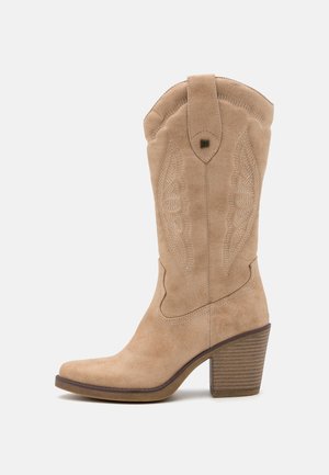 Botte cowboy mi-mollet en suède beige avec surpiqûres décoratives, languette, bout pointu et talon bloc empilé sur fond blanc.