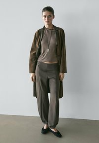 Frau trägt einen braunen Wildledermantel, passende weite Hosen, ein braunes Oberteil, schwarze Ballerinas und eine lange Halskette, steht vor einem schlichten Hintergrund.