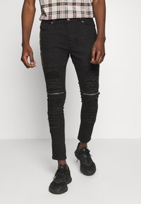 Vaqueros ajustados negros con detalles desgastados, que presentan roturas y secciones con cremalleras en las rodillas. Estilizados con zapatillas deportivas negras.