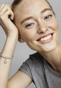 Silbernes Charm-Armband mit einem blumenförmigen Charm. Das Modell hat helle Haut, rote Haare, Sommersprossen und trägt ein graues T-Shirt. Sie hat einen lächelnden Gesichtsausdruck.