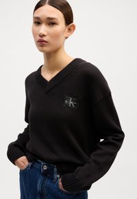 Črn pleten pulover z V-izrezom in rebrastim robom. Ima pravokotni logotip s tekstom "Calvin Klein" na levi strani.
