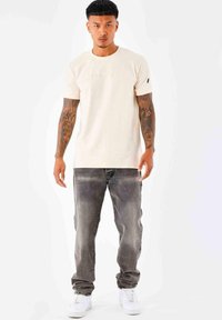 Camiseta corta de manga con textura en color beige claro, acompañada de jeans grises desgastados y zapatillas blancas, de pie contra un fondo liso.