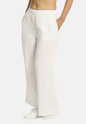 Femme portant un pantalon blanc ample à cordon avec des poches et des baskets blanches, debout devant un fond uni.