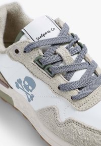 Zapatilla blanca con acentos de ante gris, hardware verde y cordones grises. Logo notable de calavera y tibias impreso en el lateral.