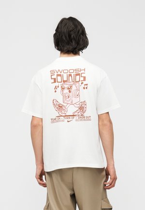 Osoba z włosami do ramion w białej koszulce z brązową grafiką przedstawiającą głośnik na nogach w sneakersach oraz tekstem „Swoosh Sounds” na plecach.