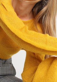 Gul mohairtröja med långa ärmar, med en mjuk, fluffig textur. Mönstrade svart-vita högmidjade byxor syns delvis.