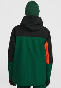 Groene en zwarte gewatteerde jas met oranje accenten. Gladde, weerbestendige stof, met een relaxte pasvorm en een verstelbare capuchon.
