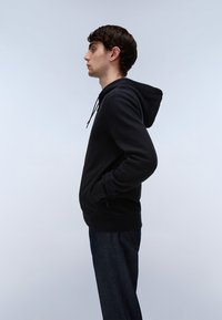 Junger Mann steht im Profil mit den Händen in den Taschen, trägt einen schwarzen Hoodie und dunkle Jeans vor einem schlichten hellen Hintergrund.