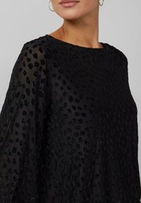 Vrouw in een doorschijnende zwarte blouse met een textuur van stippen en lange mouwen, zichtbaar van schouders tot kin met gouden ringoorbellen.