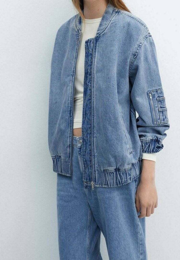 Personne portant une veste en jean bleu clair avec des poignets élastiques et un jean taille haute assorti sur un top court blanc.