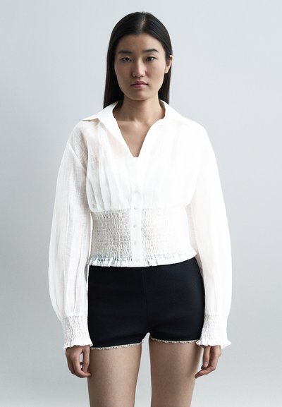 Femme aux longs cheveux foncés portant une blouse blanche à boutons avec une taille et des poignets smockés, associée à un short noir ajusté avec bordure en dentelle.