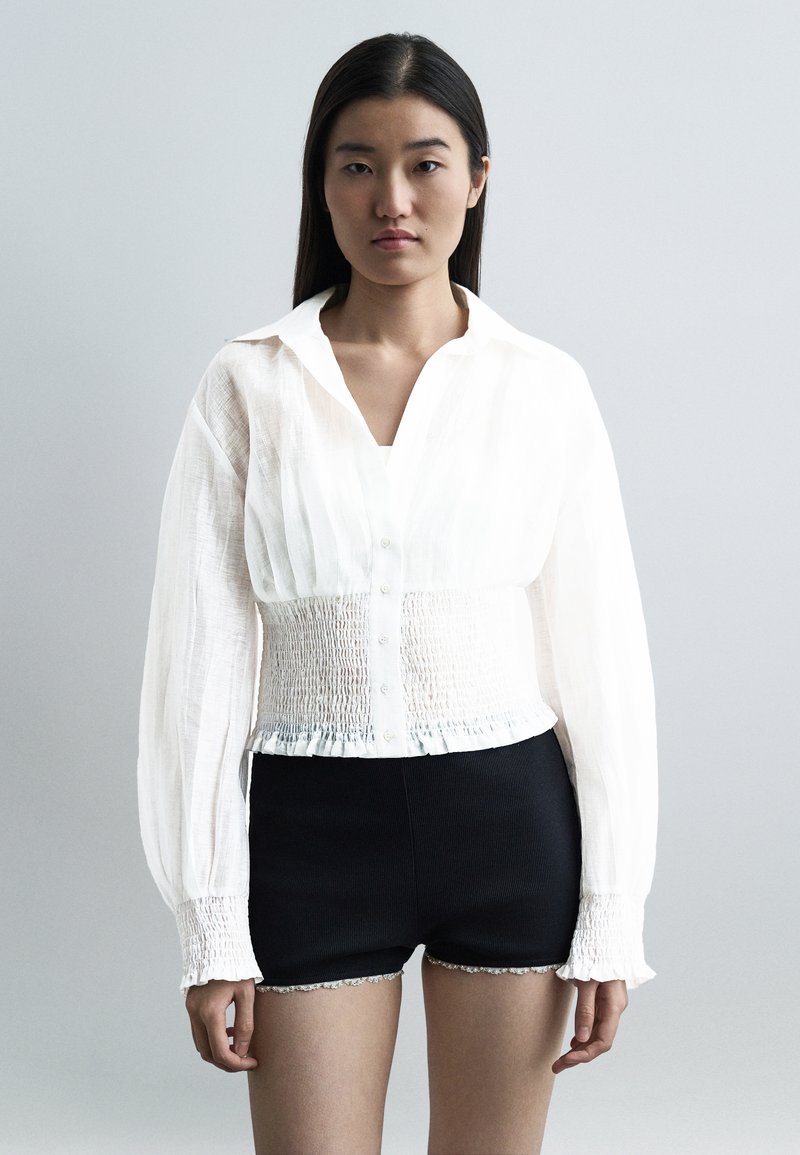 Femme aux longs cheveux foncés portant une blouse blanche à boutons avec une taille et des poignets smockés, associée à un short noir ajusté avec bordure en dentelle.