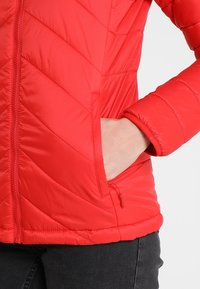 Červená péřová bunda s prošívaným vzorem ve tvaru šípu, zapínání na zip a bočními kapsami. Má elastické manžety a hladký, lesklý povrch.