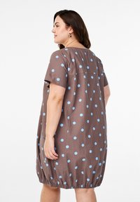 Braunes Kleid mit kurzen Ärmeln, hellblauen Punkten, abgerundetem Saum, elastischem Abschluss und einer Naht hinten. Aus einem weichen Stoff gefertigt.