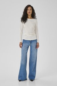 Woman with long curly hair wearing a white long-sleeve top and wide-leg blue jeans standing on a plain gray background.