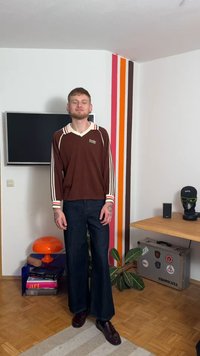 Brun langermet skjorte med kremstriper, kombinert med mørke vidtgående jeans. Skjorten har en logo, og antrekket fullføres med mørke sko. Innendørsmiljø.