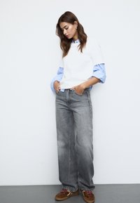 Bílý top s krátkým rukávem navrstvený přes modrou pruhovanou košili s knoflíky, doplněný šedými širokými kalhotami a hnědými nazouvacími botami.