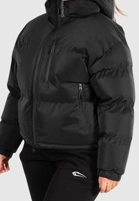 Giacca a piumino nera con collo alto, tasche con cerniera e polsini a costine, con una texture liscia e una forma squadrata.