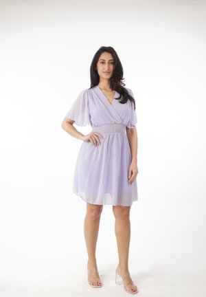 Mujer con cabello largo y oscuro que lleva un vestido lila claro hasta la rodilla y sandalias transparentes de tacón alto, de pie con una mano en la cadera.