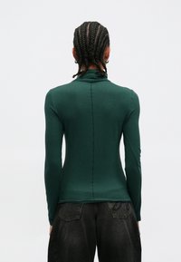 Μπλούζα με μακριά μανίκια σε σκούρο πράσινο ύφασμα, στ�ενή εφαρμογή και ψηλό γιακά. Ραφή στο πίσω μέρος. Συνδυάζεται με σκούρο γκρι παντελόνι.