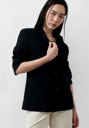 Γυναίκα με μακριά σκούρα μαλλιά που φοράει μαύρο σακάκι και λευκό παντελόνι, κρατώντας το πέτο, μπροστά σε απλό φωτεινό φόντο.