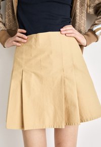 Gonna plissettata beige realizzata in tessuto di cotone, con vita piatta e una consistenza morbida. Il design enfatizza un'estetica giovanile e informale.