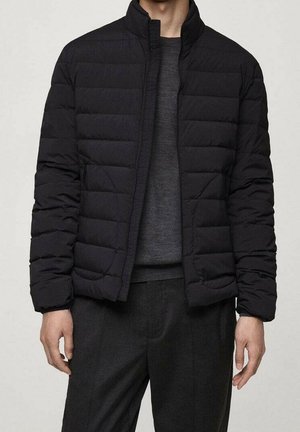 Homme portant une veste matelassée noire sur un pull gris foncé et un pantalon noir, debout avec les bras détendus le long du corps.