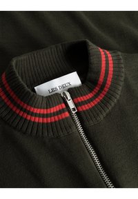 Olivgrüner Reißverschluss-Pullover mit geripptem Kragen, der rote und dunkelgraue Streifen aufweist; enthält einen silbernen Reißverschluss und ein gewebtes Etikett.