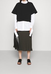 Person wearing a black cropped top over a white long-sleeve button shirt and olive pleated midi skirt, holding a black leather bag.