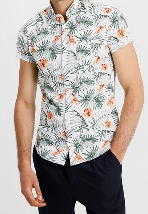Mand iført en kortærmet hvid skjorte med grønne palmeblade og orange hibiscusblomster, kombineret med sorte bukser.