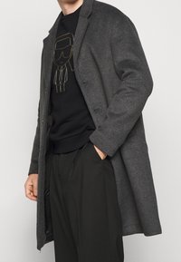 Abrigo de lana gris con cuello notch, cierre de un botón y bolsillos laterales, superpuesto a un sudadera negra con un diseño gráfico dorado.