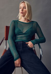 Gennemsigtig grøn top med lange ærmer og glimmer, kombineret med højtaljede sorte bukser, siddende på en stol af metal og træ, med afslappede arme.