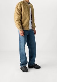 Muž v béžové bombr jakně, bílé košili, modrých džínech a černých teniskách stojí před bílým pozadím.