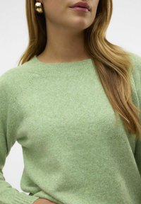 Pull vert clair en tissu tricoté doux et texturé, avec un col rond et des manches raglan. Remarquable pour sa variation subtile de couleur.