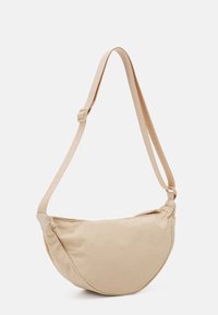 Beige půlměsícová crossbody taška s nastavitelným textilním popruhem a horním zipovým zavíráním na bílém pozadí.