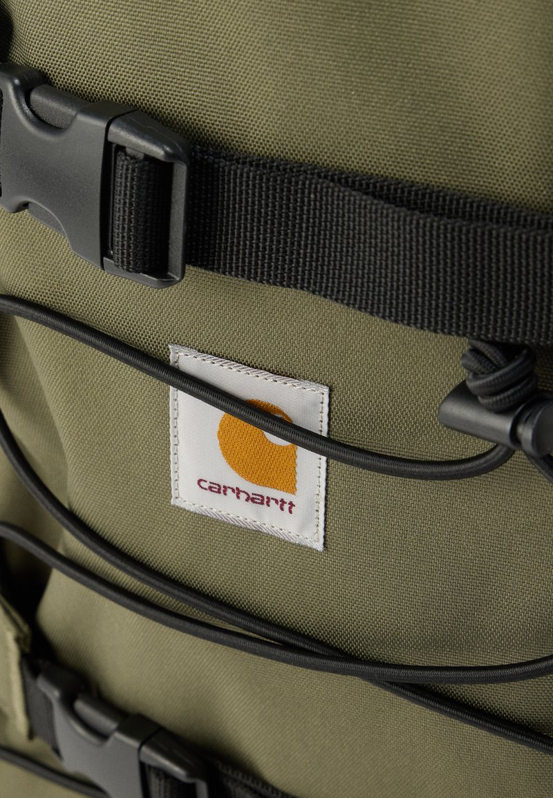 Olivgrüner Rucksack mit strukturiertem Stoff, schwarzen Gurten, Kunststoffschnallen und einem weißen Etikett mit gelbem Logo.