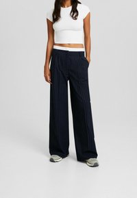 Marineblauwe pinstripe wijde pantalon met een witte elastische tailleband; gedragen met een strakke witte cropped top en grijze sneakers. Gladde stofstructuur.