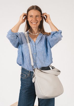 Smiling woman adjusting hair, wearing blue striped shirt, jeans, shell necklace, and carrying large white crossbody leather bag.