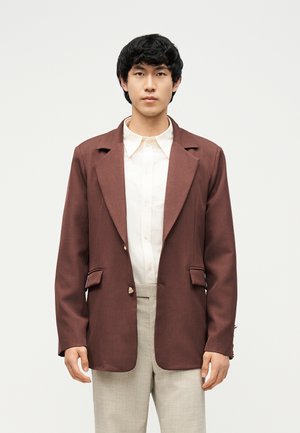 Noor mees kannab pruuni bleiserit valge nööbitava särgi ja heledate beežide pükstega, ta seisab tühja heleda tausta ees.