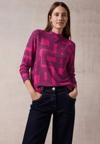 Fuchsia gebreide trui met geometrisch zwart patroon, hoge halslijn en lange mouwen, gecombineerd met donkerdenim jeans met zichtbare stiksels.